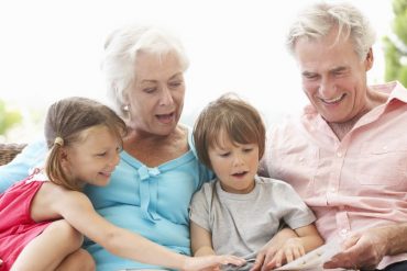 Grandparents And Grandchildren Reading Book On Garden Seat